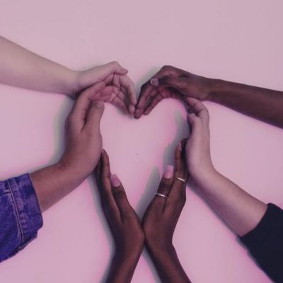 Six hands of people of various races, shaped together forming a heart on white background; purplish light over whole image