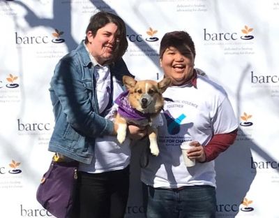 Two people holding a corgi in a BARCC bandana against a backdrop with the BARCC logo on it