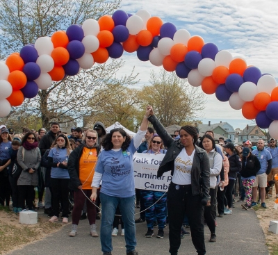 Gina Scaramella and Ayanna Pressley raise hands together in front of Walk with balloon arch above.