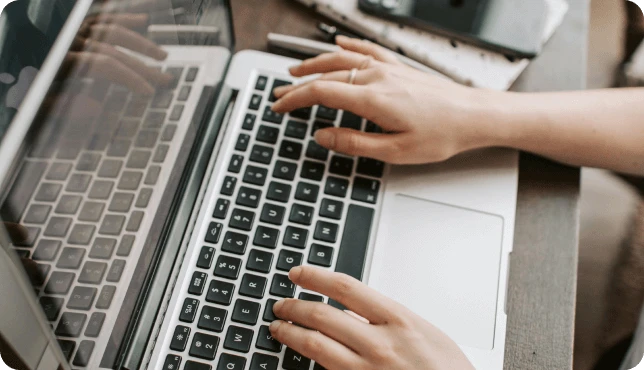 Manos escribiendo en el teclado de una computadora portátil, con un teléfono inteligente y una computadora portátil cerca.
