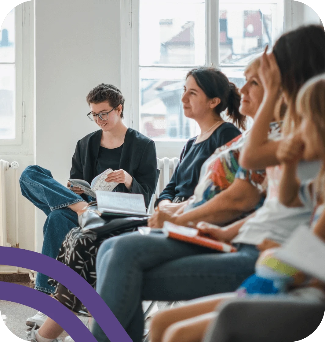 A group of people sit in chairs, some holding notebooks, in a light-filled room with large windows.