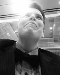 Cory-at-Gala Black and white photo of a person in a suit and bow tie looking upward, with a bright light above them.