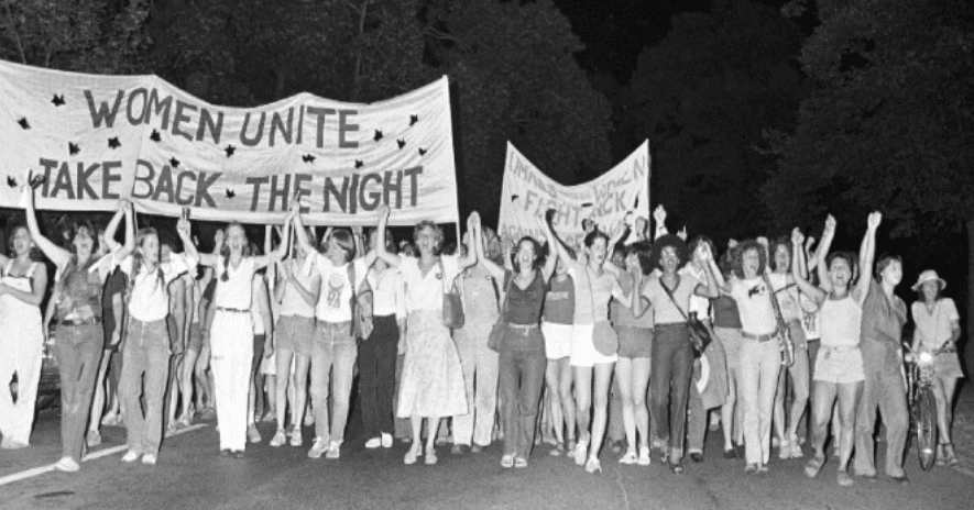 1972-barcc-founding A historic gathering unfolds as a large group of women march at night, holding banners that say "Women Unite" and "Take Back the Night.