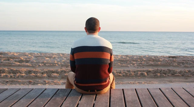 1998-male-survivor A historic scene unfolds as a person sits on a wooden deck facing the beach, overlooking the sea under a clear sky.