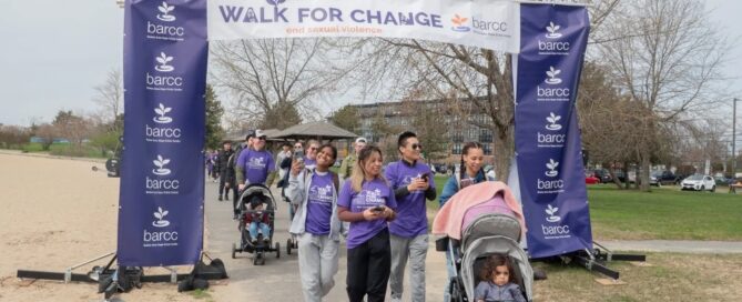 Participants in purple shirts walk under a "Walk for Change" banner, pushing strollers on a paved path. This Boston event supports the local rape crisis center, encouraging everyone to get help and raise awareness.