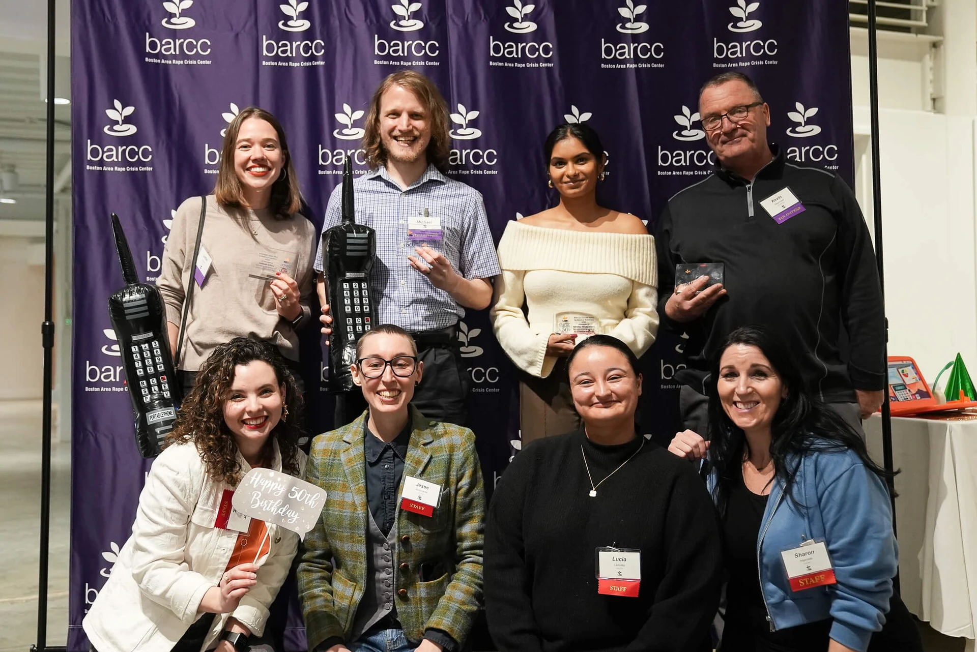 A group of nine people proudly pose in front of a "barcc" banner, some holding awards and props, highlighting the vital services offered by this rape crisis center.