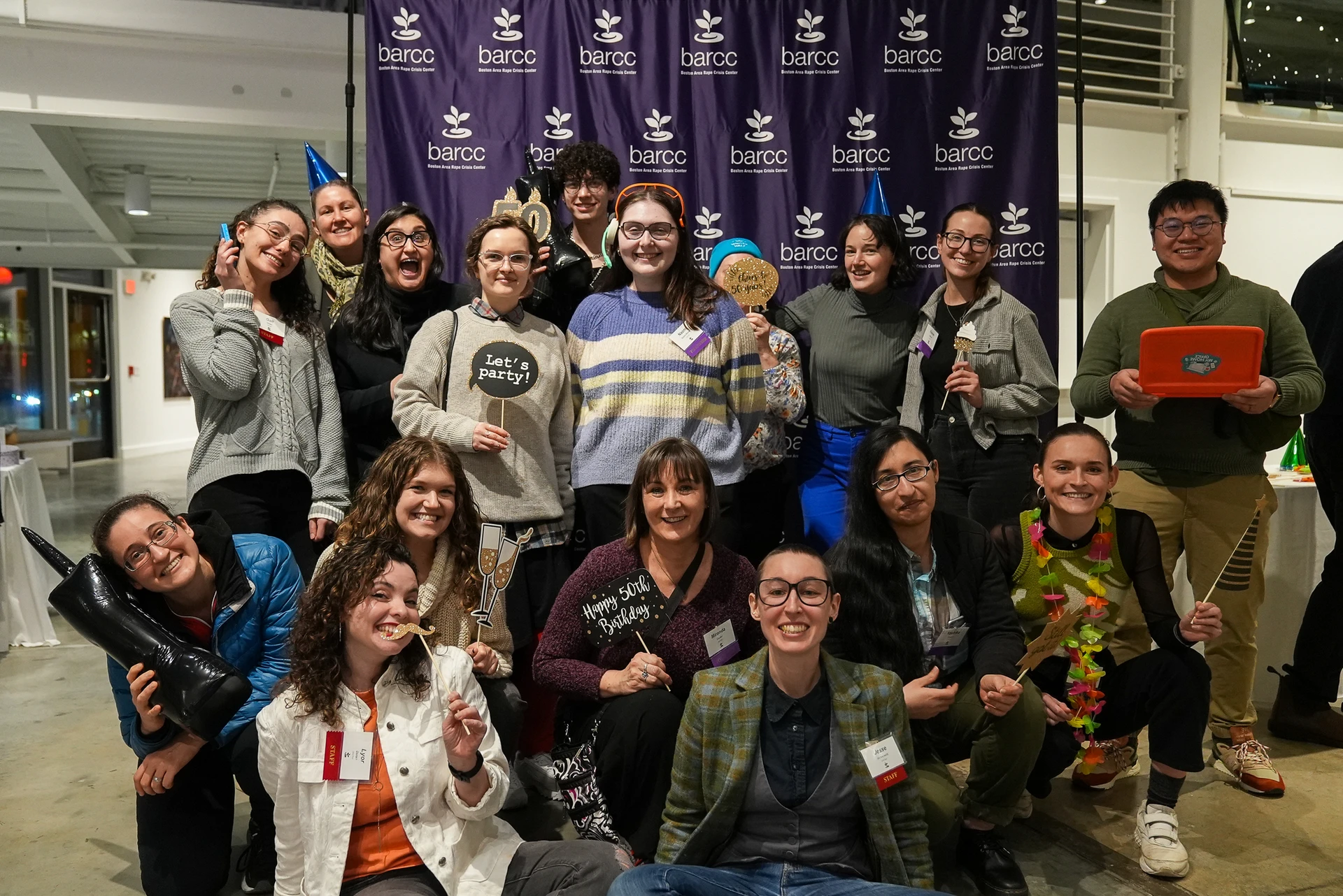 A7S01912 A group of 15 people pose playfully in front of a backdrop featuring the BARCC logo, holding various props and expressions at an indoor event.