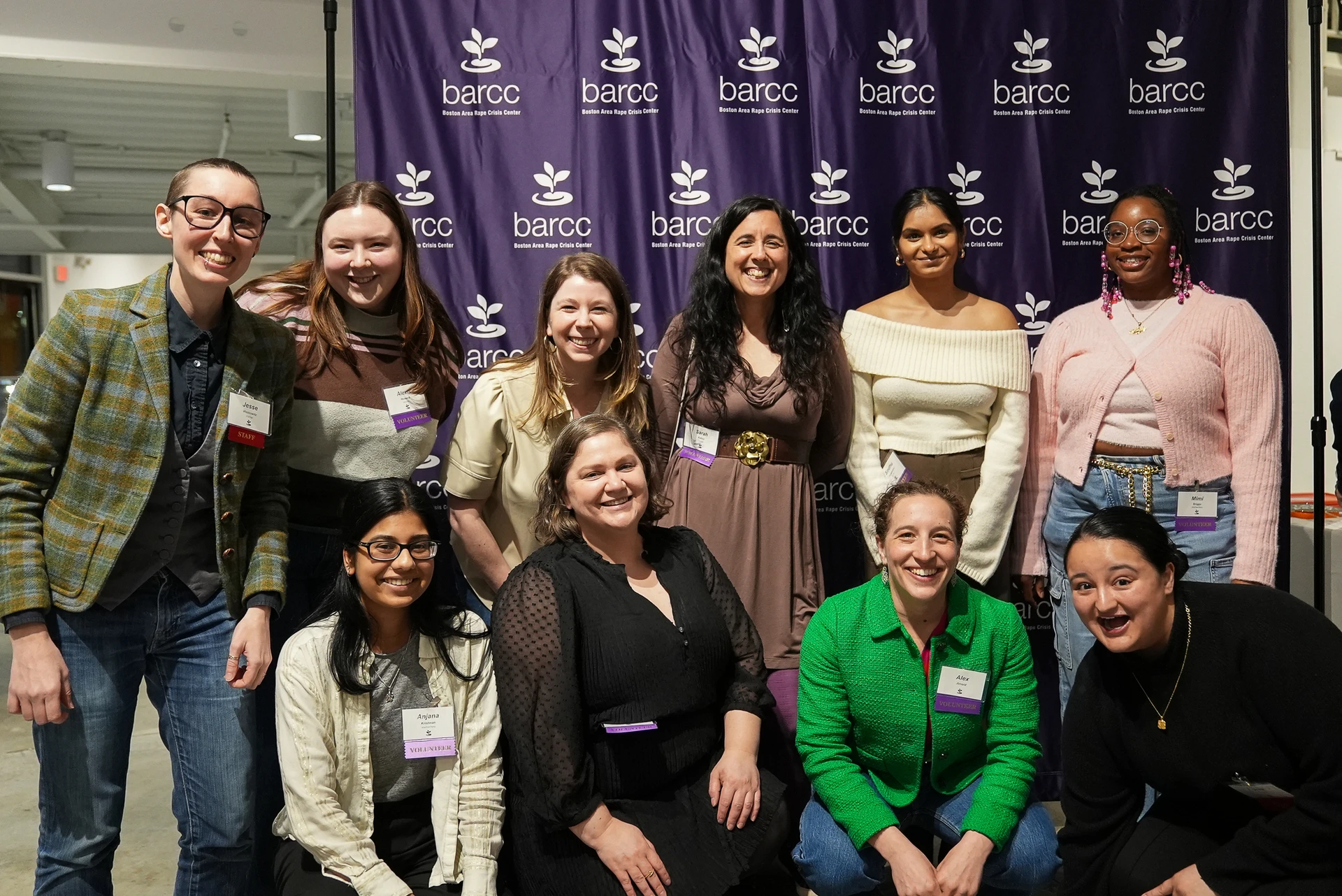 A7S01917 A group of ten people is smiling and posing indoors in front of a vibrant purple BARCC banner, radiating joy and camaraderie.