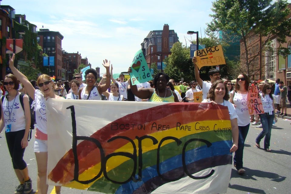 BARCC_pride People marching in a parade hold a banner for the Boston Area Rape Crisis Center, with colorful shirts and signs expressing pride. Among them, Queer Survivors stand strong, showcasing resilience and unity.