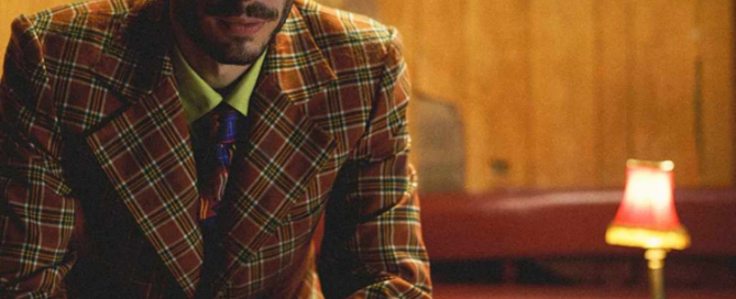 A person in a checkered suit sits in a dimly lit room with wood-paneled walls and a small lamp on a table, pondering newfound ways to offer services and get help for those in need.