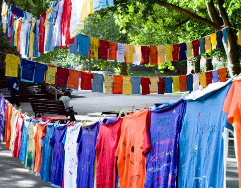 Colorful T-shirts with messages hang on clotheslines in a park, surrounded by trees, with a bench in the background.
