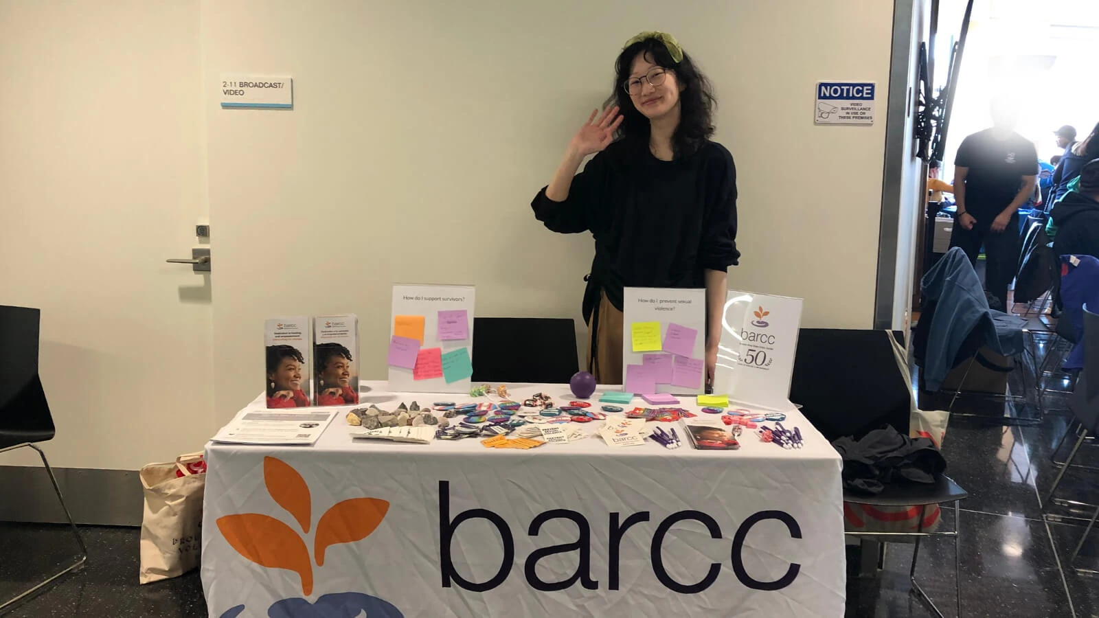 Education and Prevention A person stands behind a table adorned with brochures, buttons, and a banner reading "barcc," dedicated to education and prevention of sexual violence. They're smiling and waving inside the building, inviting engagement.