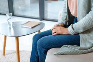 Persona sentada en un sofá con las manos entrelazadas, vestida con un suéter y jeans. Cerca de una mesa pequeña hay un cuaderno y un vaso de agua.