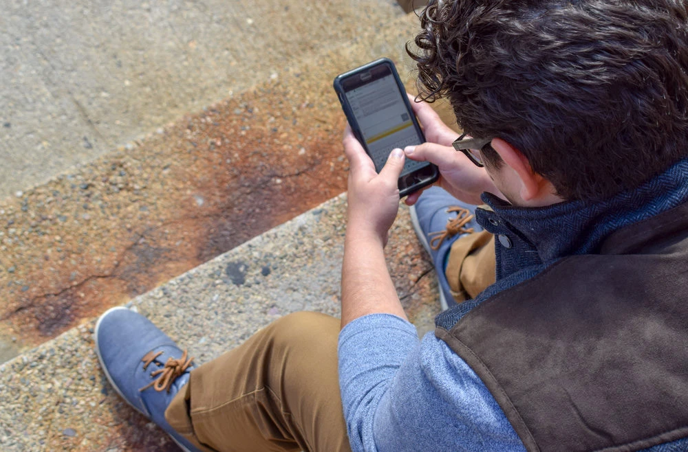 Hotline-BARCC A person with curly hair sits on the concrete steps, focused on their smartphone. They're sporting a blue long-sleeve shirt, brown pants, gray shoes, and a brown vest. Lost in thought, they consider whether to Call BARCC Now for support.