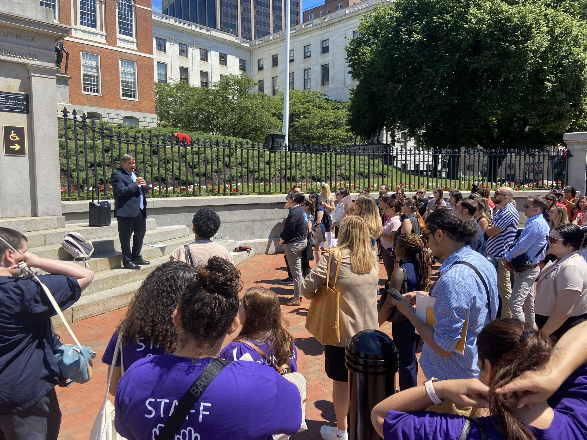 IMG_4107 A man addresses a crowd gathered outside a large building with a black fence and brick path, near the BARCC office.