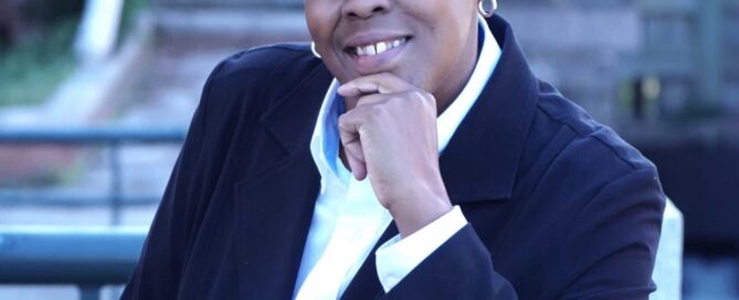 A person in a dark suit and white shirt smiles, resting their chin on their hand outdoors with a blurred brick and railing background, perhaps reflecting the historic charm of Boston.