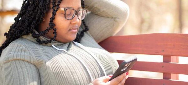 A person with glasses is sitting on a wooden swing in Boston, looking at a smartphone, with one hand behind their head. They are wearing a gray sweater, perhaps researching local services.