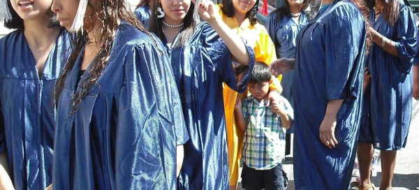 A group of Boston graduates in blue gowns and caps, along with one in a yellow outfit, stand outdoors in line. A child stands among them holding an adult's hand, symbolizing the supportive services available to help families celebrate this milestone together.