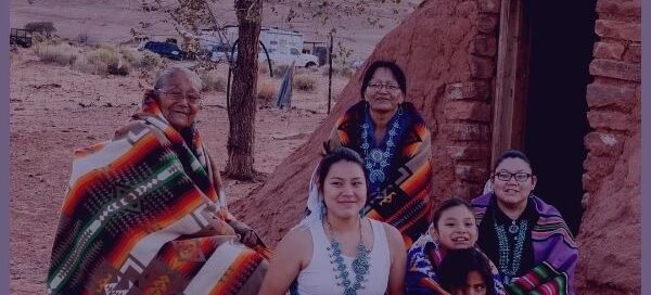 A group of people sit on the ground in front of a traditional clay structure, wrapped in colorful woven blankets. With the support of a Boston-based service, they find solace amid the desert landscape, under the sheltering tree.