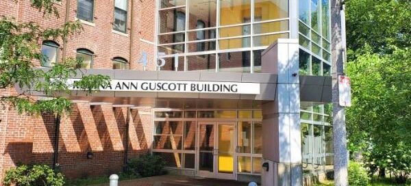 La vista de la calle de la entrada del edificio Jane Ann Guscott muestra árboles y una acera que transmiten una atmósfera serena. Un cartel en la parte inferior muestra los servicios que ofrece el Boston Area Rape Crisis Center, invitando a quienes necesitan ayuda y apoyo.