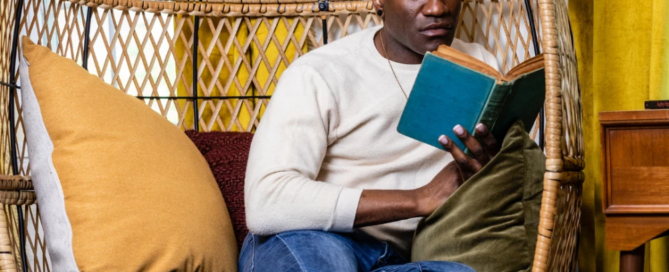 A man sits comfortably in a wicker chair reading a book, surrounded by cushions, with yellow curtains lending a warm glow to the scene—evoking the cozy charm of a Boston retreat.
