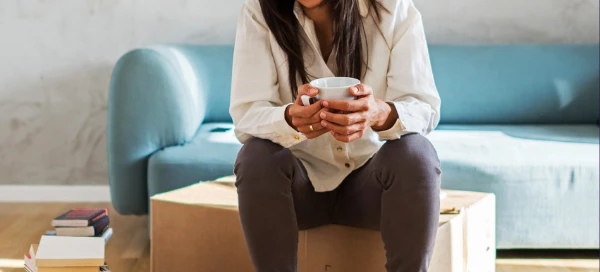 A woman sits on a cardboard box, holding a mug, with a stack of books nearby and a blue sofa in the background. In this cozy Boston setting, she finds comfort in her surroundings and gets help from the stories within those pages.