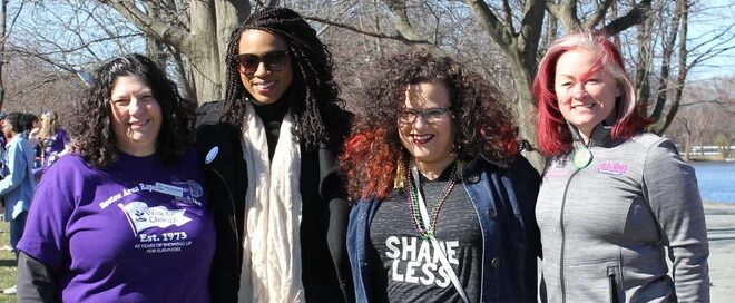 Four people standing outdoors, smiling, with trees and a body of water in the background, embody the spirit of unity and support found at Boston's dedicated services like the rape crisis center.