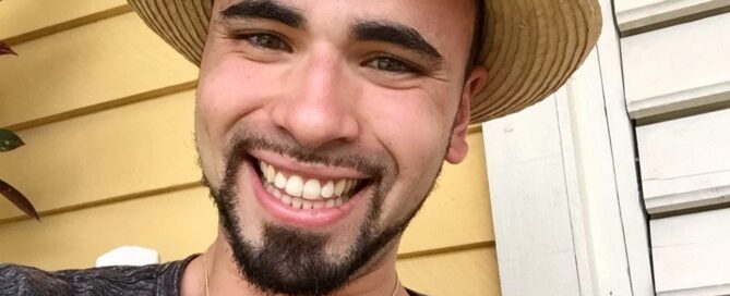 A man smiling outdoors in a straw hat and gray shirt, sitting against a yellow wall with white window shutters, radiates warmth and tranquility—a reflection of the compassionate services one might find when seeking to get help at a local center.