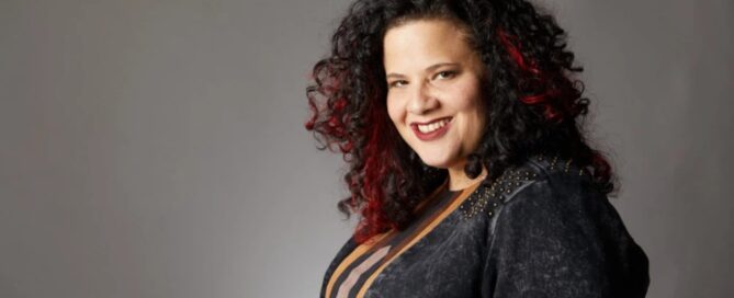 A person with curly hair and a smile, donning a dark outfit with vertical stripes, stands poised against a gray background, ready to offer their services.