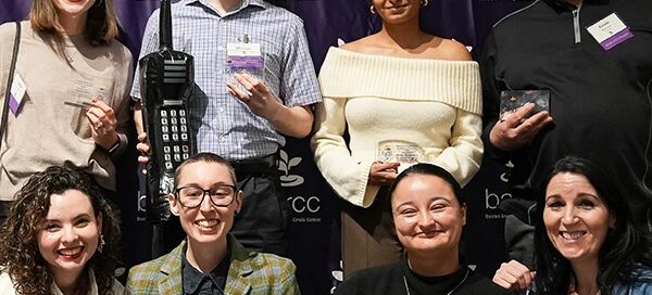 A group of people smiling and posing in front of a "barcc" banner, some holding award items and badges, celebrating the Boston Rape Crisis Center's efforts to support those seeking help.