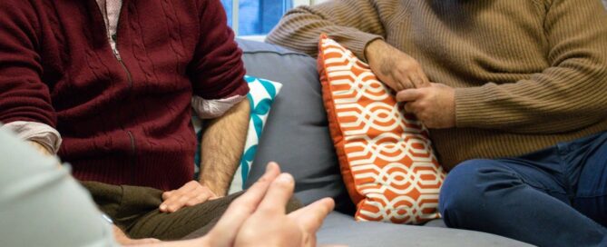Three people seated on a couch engaged in conversation, their body language and hands conveying comfort and support, surrounded by colorful cushions. It's a scene emphasizing connection, reminiscent of the supportive environment found in a crisis center where individuals can get help.