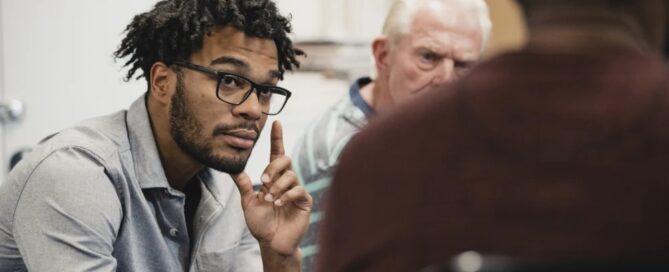 In a Boston-based discussion, a man wearing glasses attentively listens, embodying the supportive atmosphere encouraged by local centers that help individuals in need. Two others are partially visible in the background, contributing to this meaningful exchange.