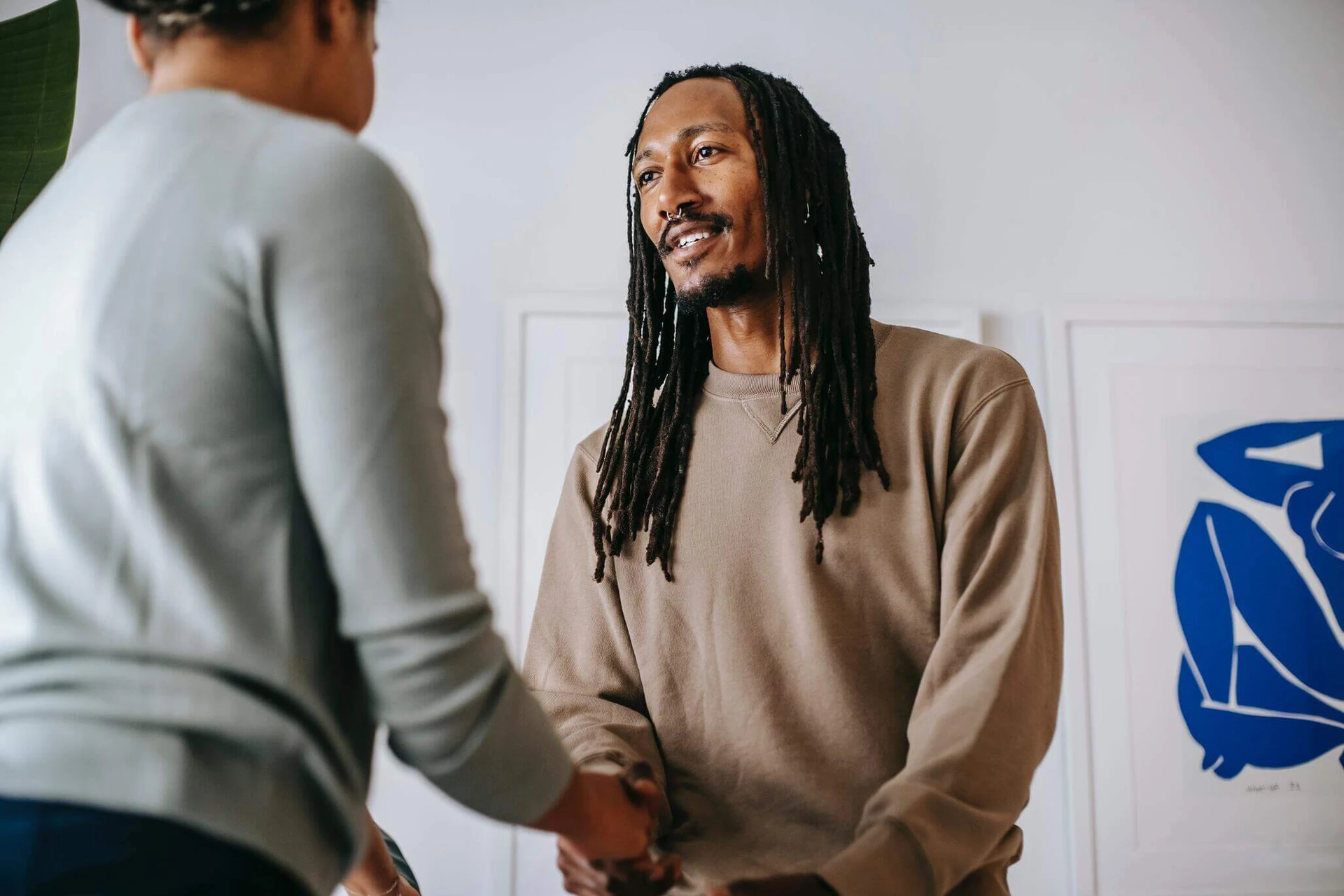 Two people are shaking hands in a room adorned with framed artwork. One, wearing a brown sweatshirt, has long hair; the other sports a light gray top. The meeting takes place in Boston's community hub, where essential services like the local rape crisis center are just around the corner.