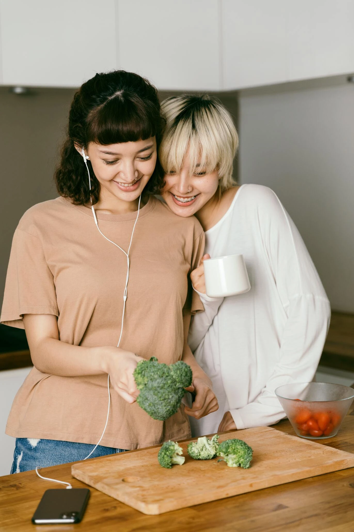 Two people are in a kitchen, practicing self-care. One holds a piece of broccoli with earbuds in, while the other leans close with a white mug. A bowl of tomatoes and a phone lie on the counter, creating a serene space for coping exercises and connection.
