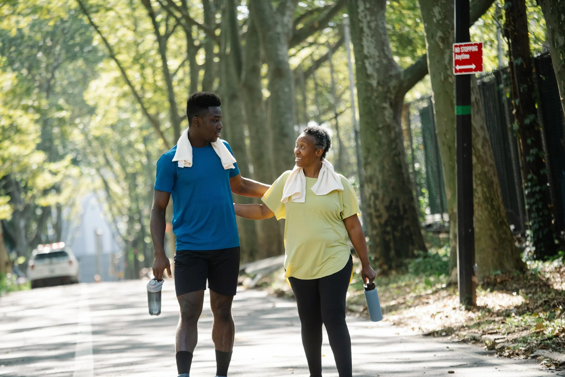 pexels-liliana-drew-8497748 Two people walking on a tree-lined road with towels around their necks and water bottles in hand, engaged in conversation, as they support each other to get help.