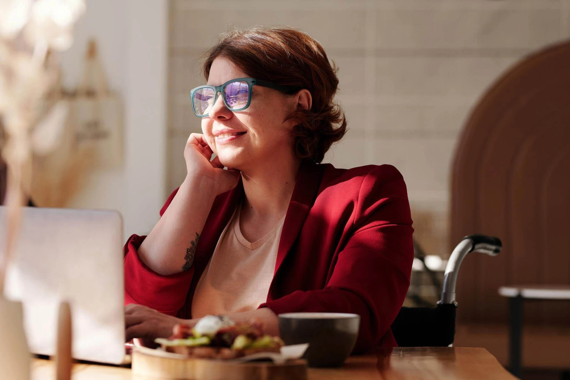 A woman in glasses and a red blazer sits at a table with a laptop and food, smiling in natural sunlight as she researches services at the local rape crisis center.
