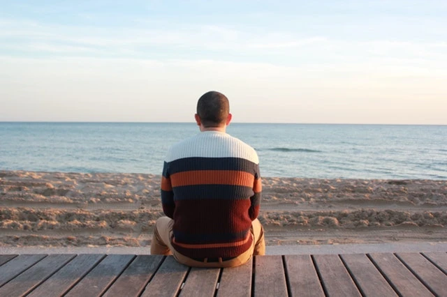 pexels-photo-207920 A male survivor in a striped sweater sits on a wooden deck, gazing at the tranquil ocean under a clear sky.