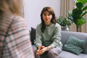 Una mujer con un suéter verde está sentada en un sofá gris y escucha atentamente. Una persona con cabello largo, de espaldas a la cámara, también está sentada en el sofá. Al fondo hay una planta en maceta.
