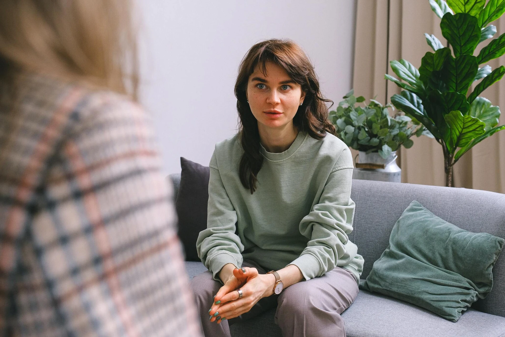 A woman in a green sweatshirt sits on a gray couch, talking to another person. Green plants are in the background, creating a calm atmosphere as they discuss how to get help from a local rape crisis center in Boston.