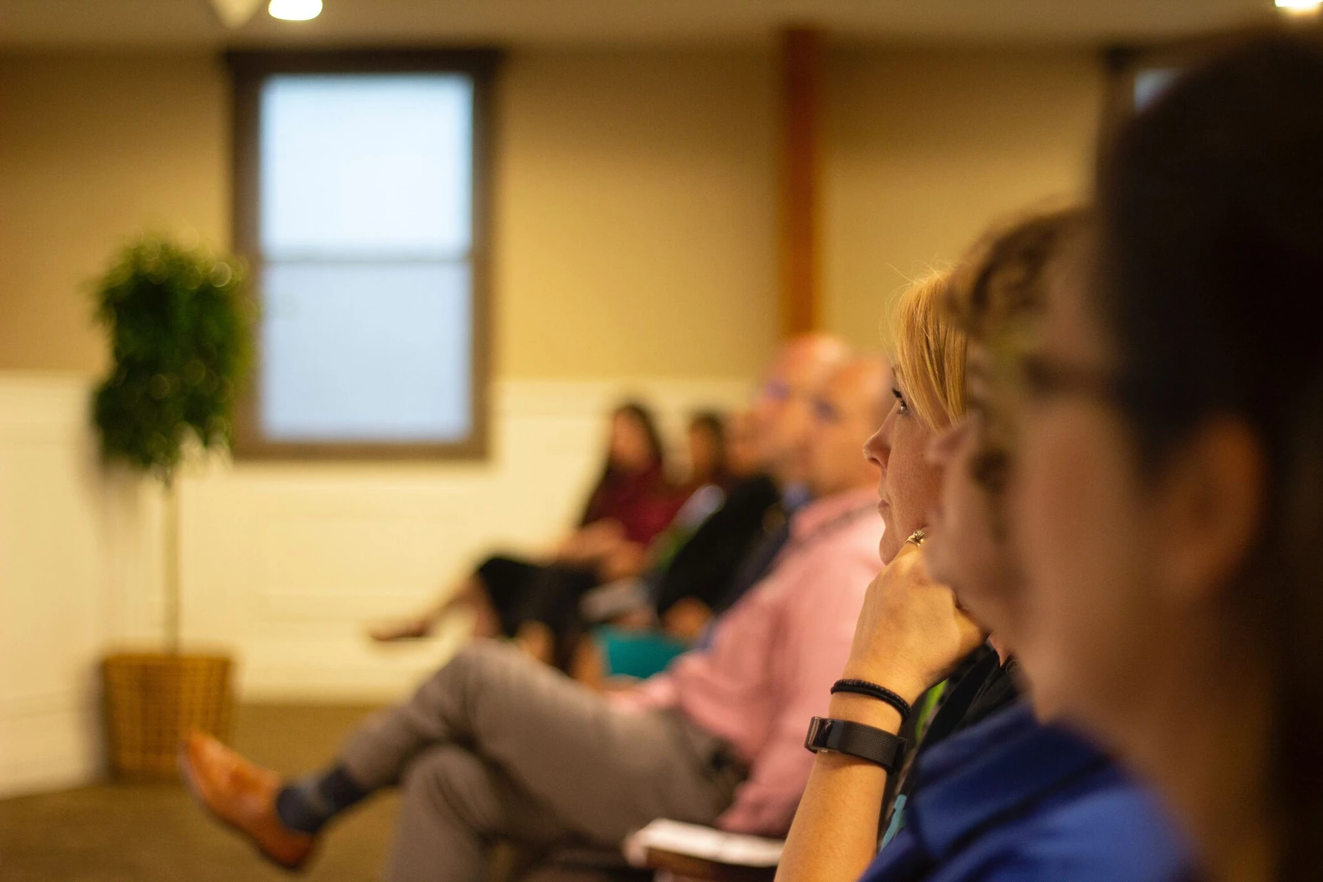In a softly lit room in Boston, people are seated attentively, listening as they get help. A plant and a window provide a serene backdrop.