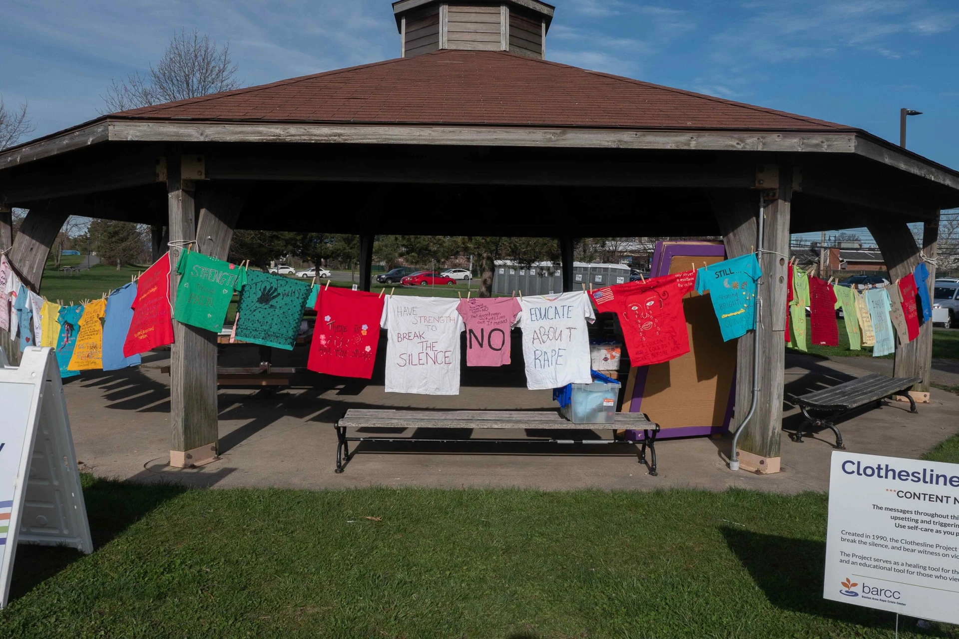 A Boston gazebo showcases colorful shirts with messages about breaking silence and educating against violence, urging the community to get involved as part of a public awareness display.