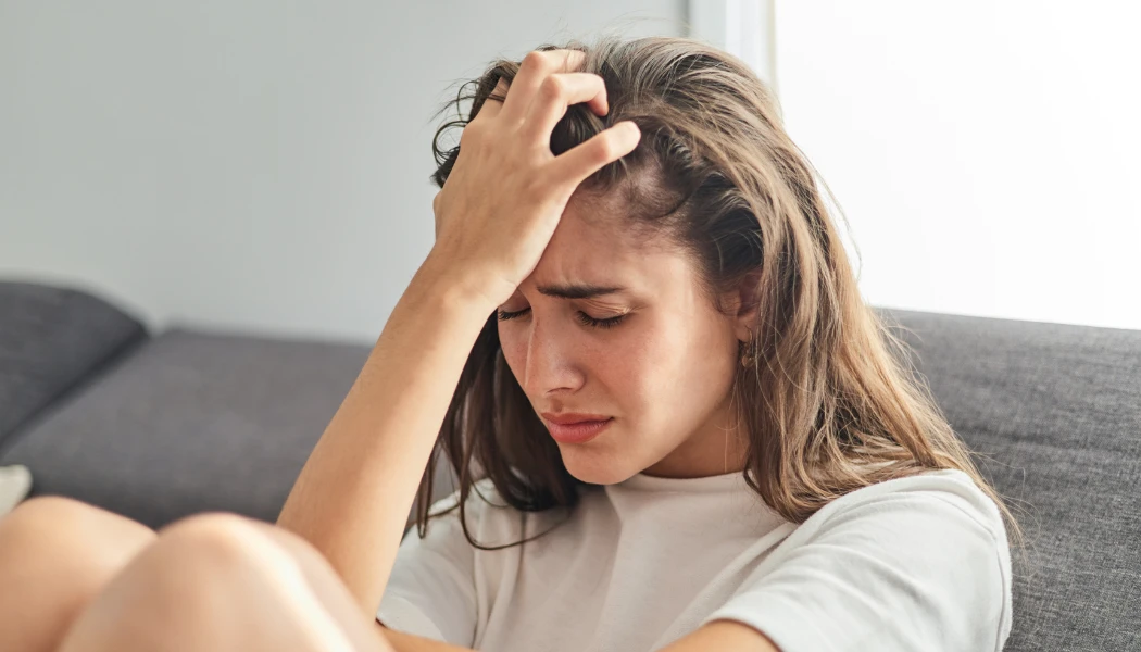BARCC Get Help Tile A woman with long hair sits on a couch, wearing a white shirt, lost in thought as if pondering life's frequently asked questions.
