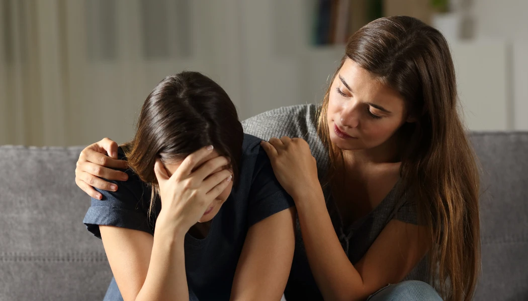 Una mujer se sienta en un sofá, sosteniendo su cabeza entre sus manos, mientras otra mujer se sienta a su lado y le ofrece consuelo con una mano en su hombro.