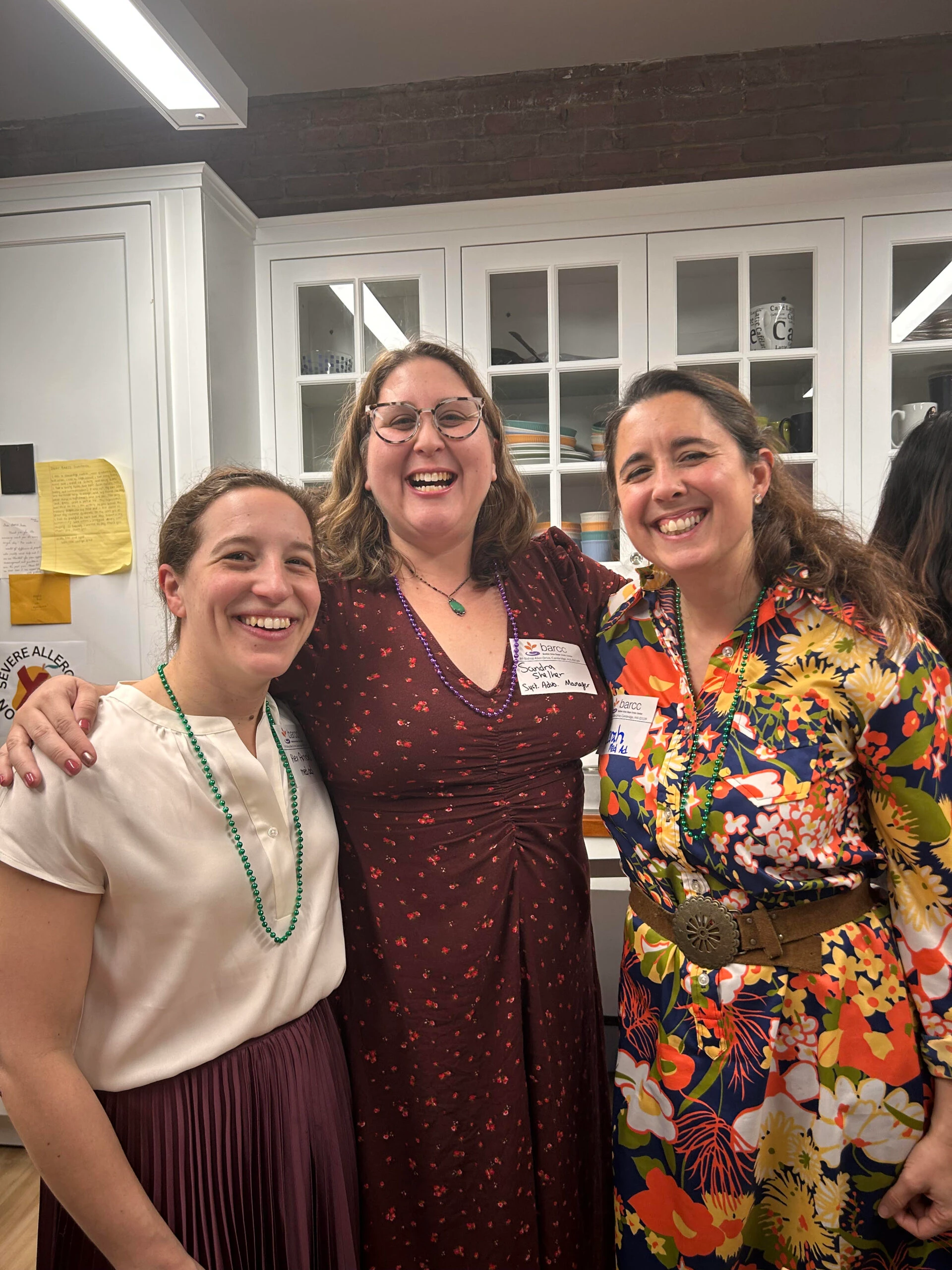 Three people, all smiles, stand together in a kitchen, with sleek cabinets and a polished countertop in the background, creating a warm and inviting scene reminiscent of a lively BARCC community gathering.