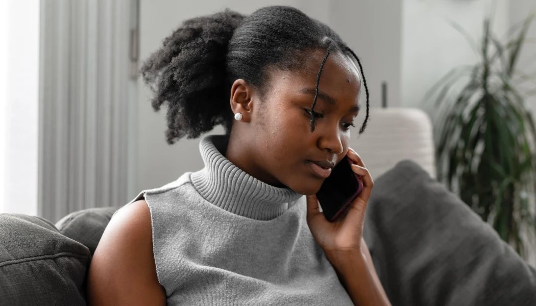 A woman in a gray sleeveless turtleneck sits on a couch, holding a smartphone to her ear, possibly seeking to get involved with or get help from a rape crisis center. A plant is visible in the background.