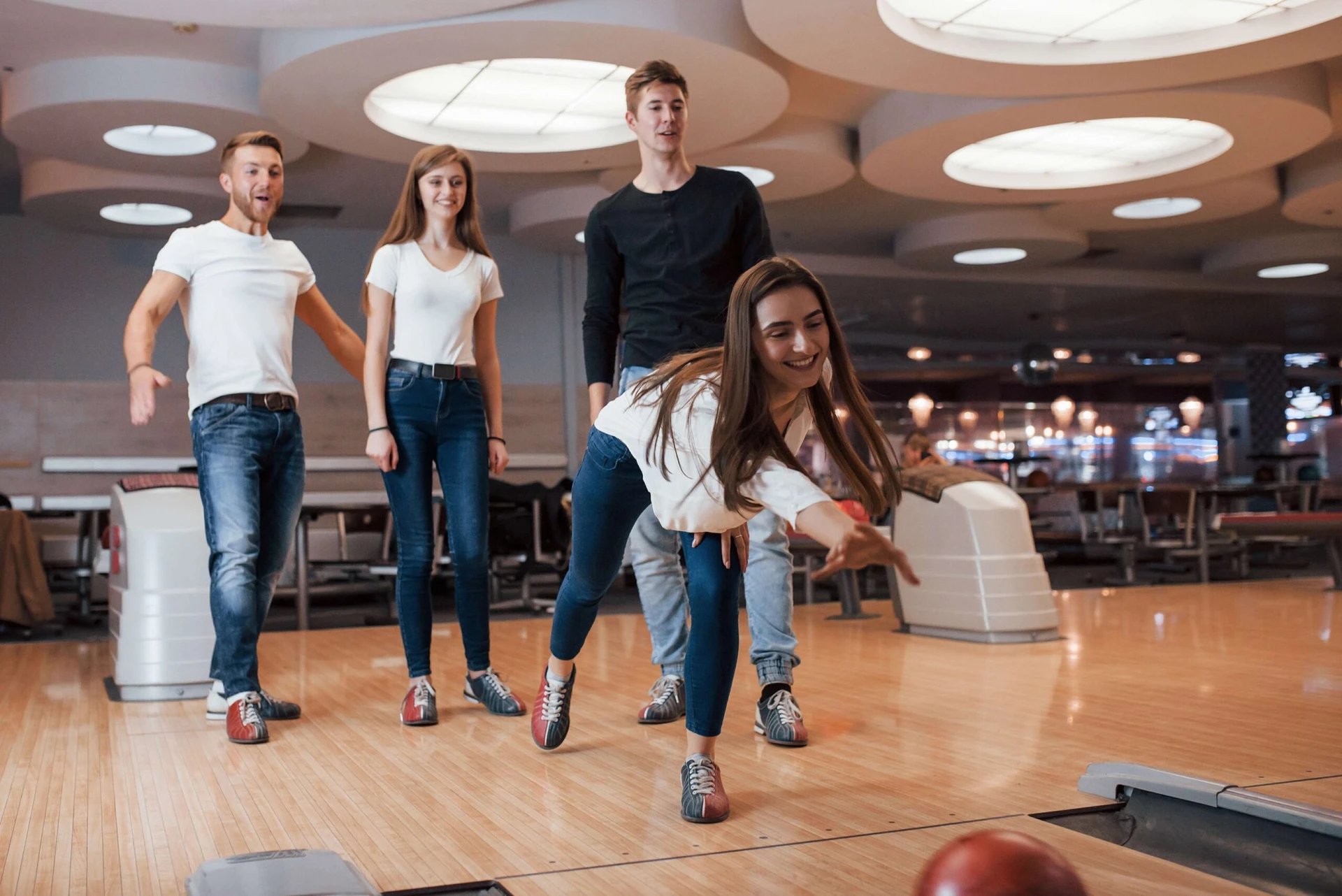 Four young adults are bowling. One person is in the process of releasing the ball down the lane while the others watch. The scene is set in a modern bowling alley.