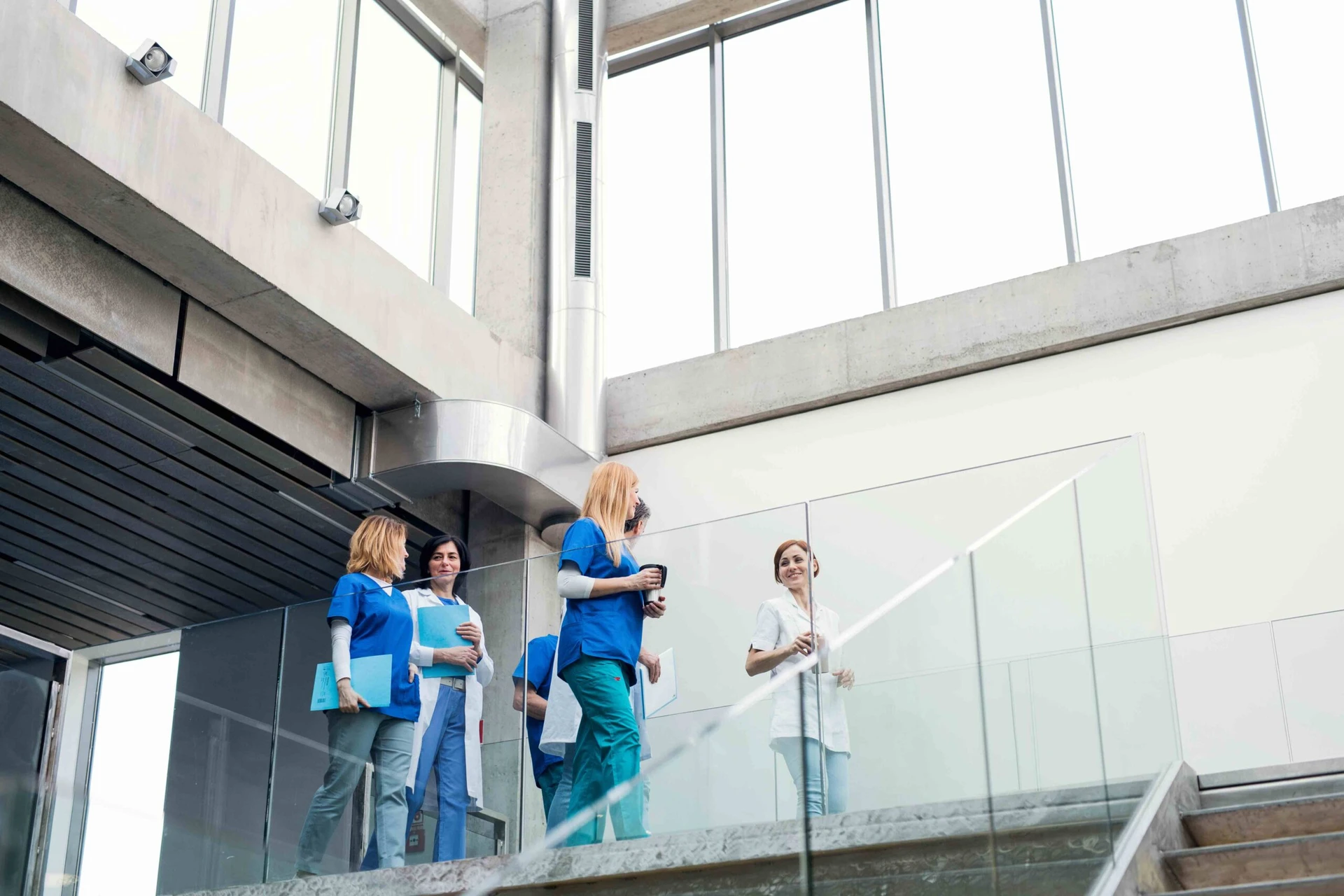 A group of dedicated medical professionals in blue and white coats, including a volunteer eager to contribute, walk down a modern building's staircase, holding files and cups.