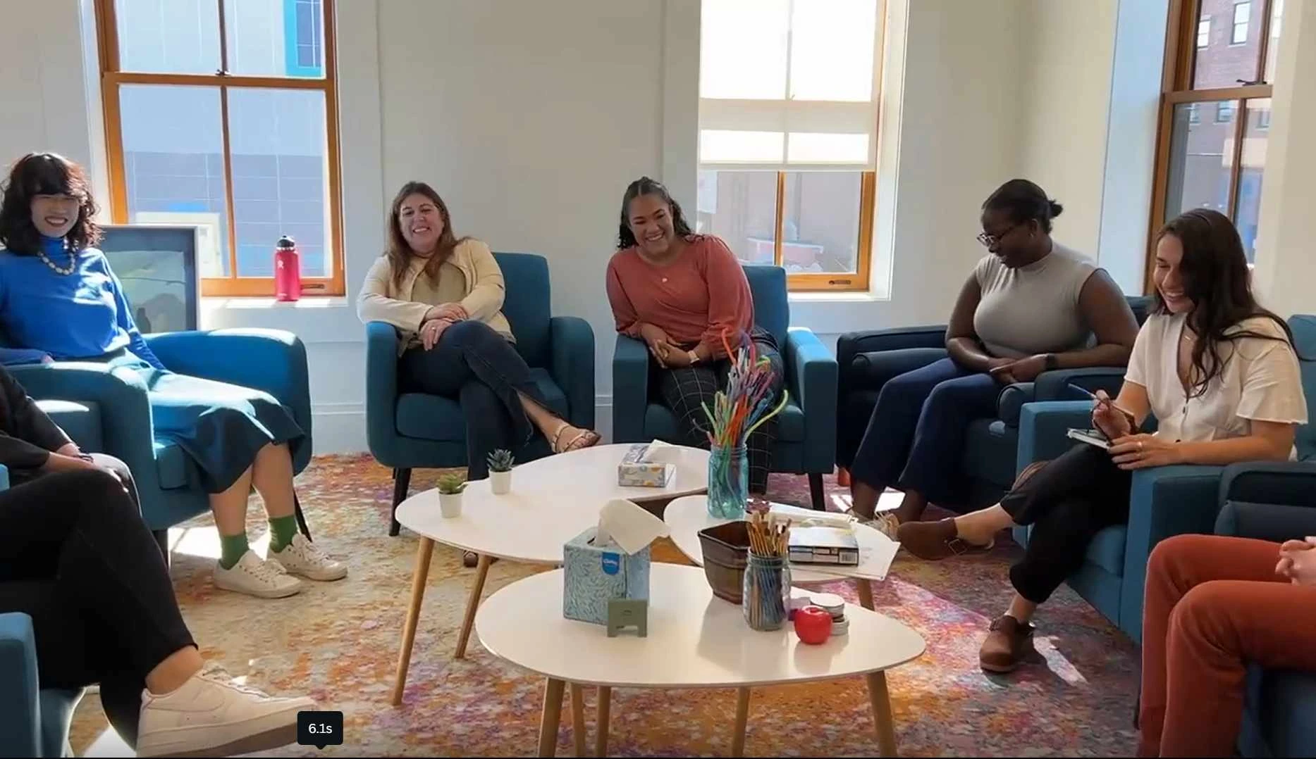Six people are seated in a circle of blue chairs in a well-lit room with large windows, engaged in conversation. A round coffee table with various items, including a booklet of Frequently Asked Questions, sits at the center.