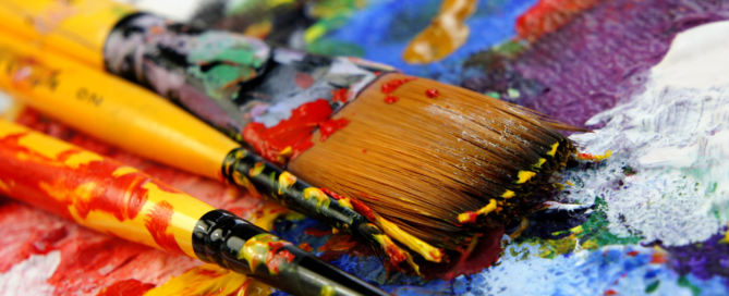 A close-up of paintbrushes covered in colorful paint rests on a vibrant, multi-colored palette, embodying the mission of barcc.org to create something beautiful and new with every stroke.