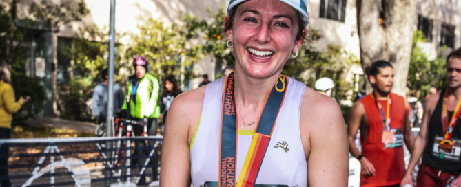Emily Mellen wearing a medal and holding a water bottle smiles triumphantly after the Boston Marathon, standing in front of a barrier with others celebrating in the background.